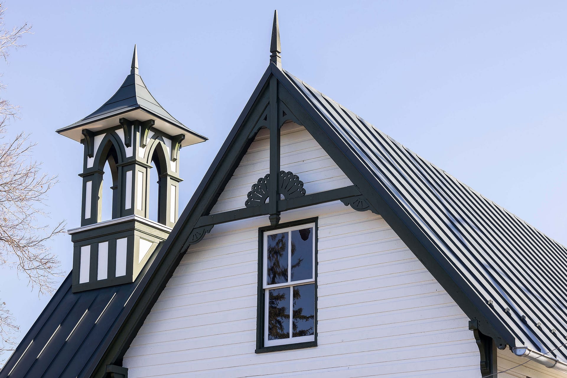 The image shows the upper section of the Elmwood Cemetery's superintendents house in Shepherdstown, WV. It has dark trim, a steep metal roof, a central decorative gable and a ornate bell tower on the left side. Originally built in 1881, restored with Ottosson linseed oil paint by Vintage Renovation and Construction.